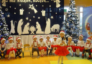 Dziewczynka recytuje wiersz, trzyma w ręku mikrofon w tle dzieci siedzą na krzesłach na tle dekoracji świątecznej, choinek.